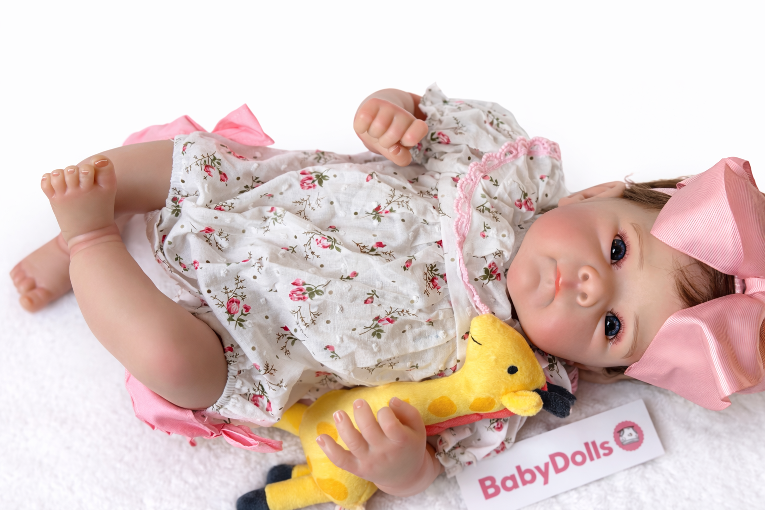 Baby doll wearing a floral outfit with a pink bow, holding a yellow toy, on a white background with 'BabyDolls' branding.